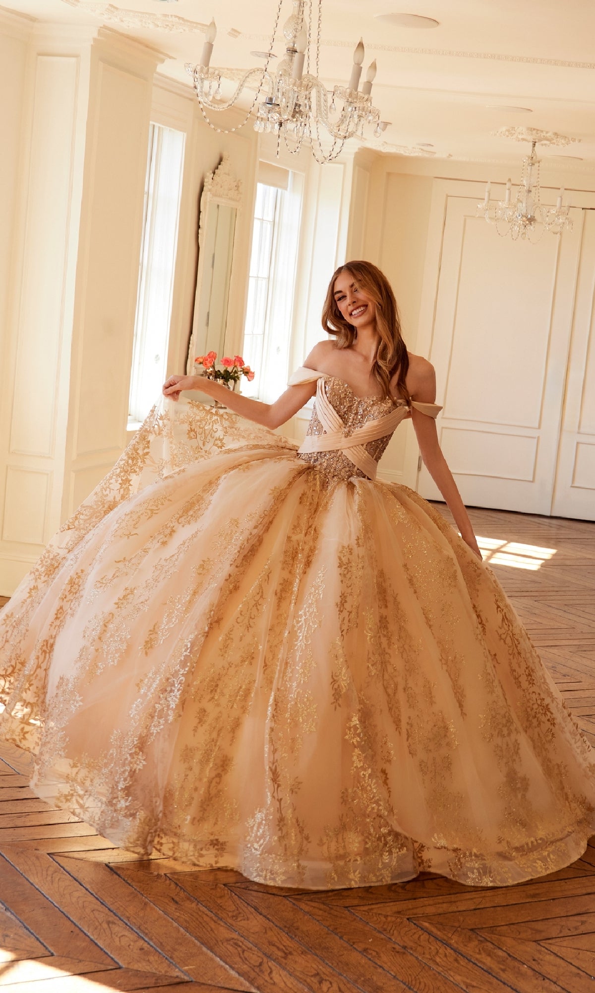 In a spacious, elegant room, a woman smiles as she holds out the skirt of the Quinceanera Dress JT1488H By Juliet, featuring gold embellishments, a ball gown skirt, and corset bodice. A sparkling chandelier and flowers decorate the background.